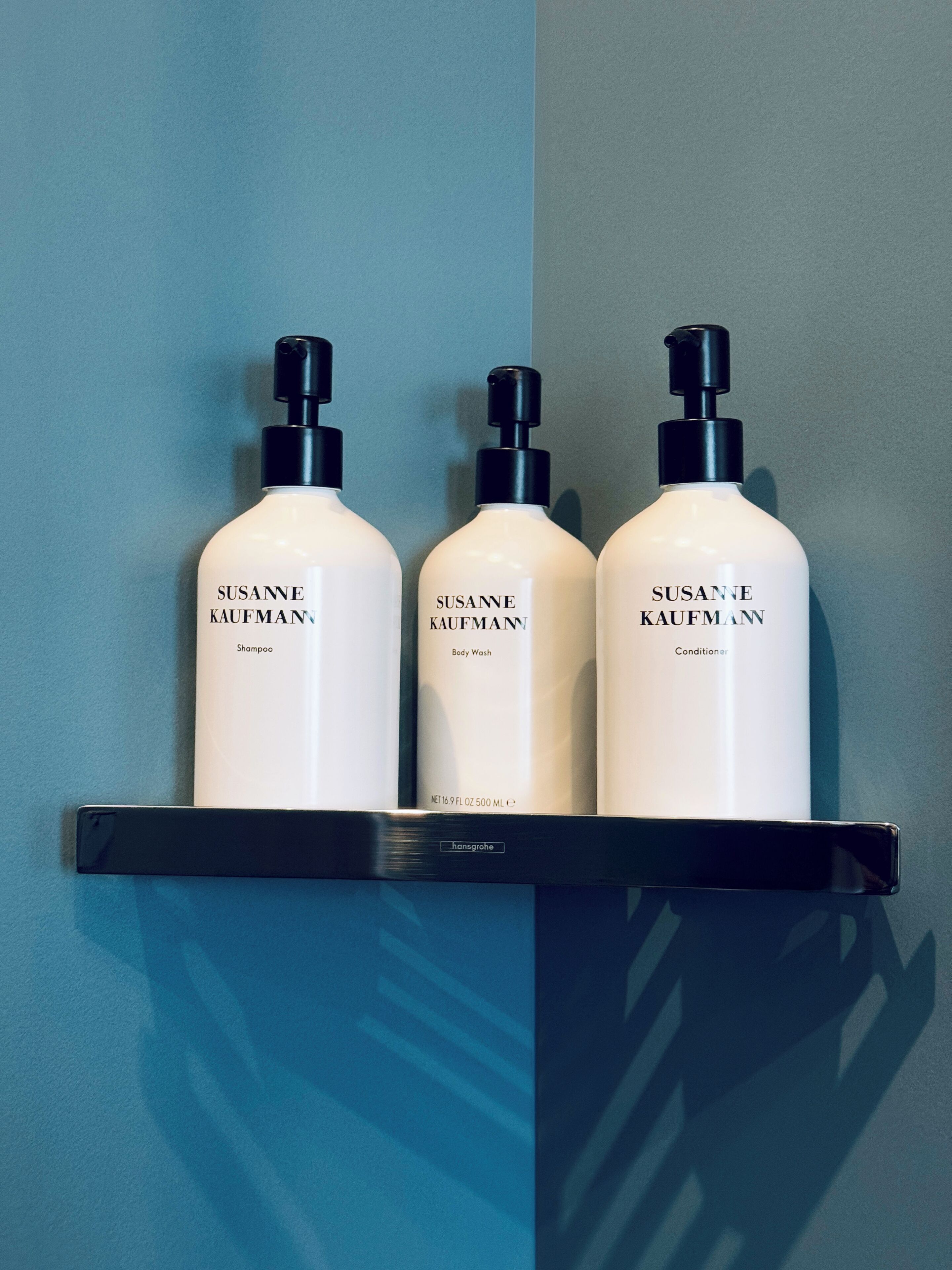 Three bottles of Susanne Kaufmann products on a black shelf against a blue wall.