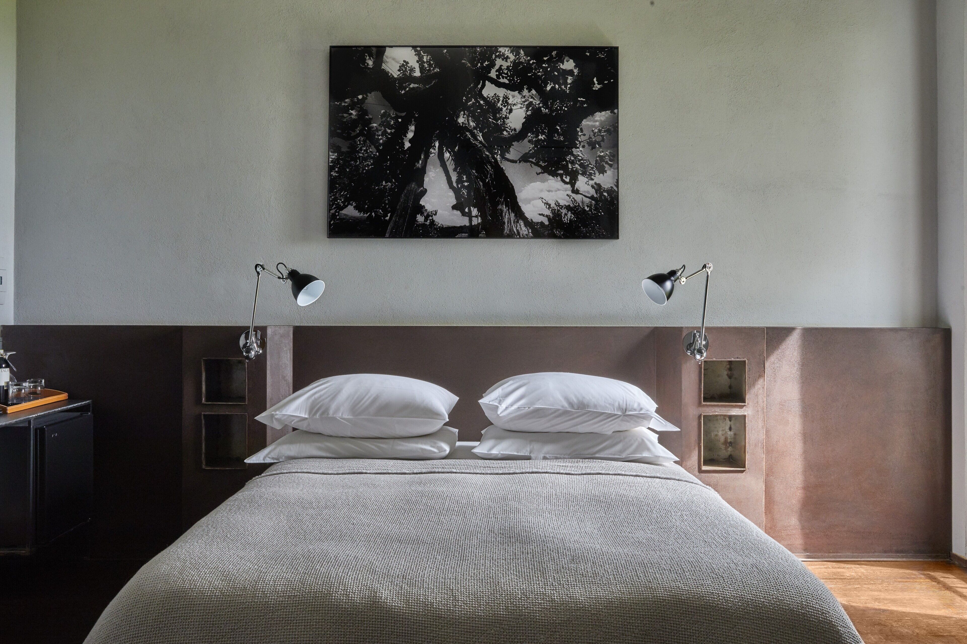 Ein ordentlich bezogenes Bett mit weißer Bettwäsche, zwei Nachttischlampen und ein gerahmtes Bild von einem Baum an der Wand darüber.