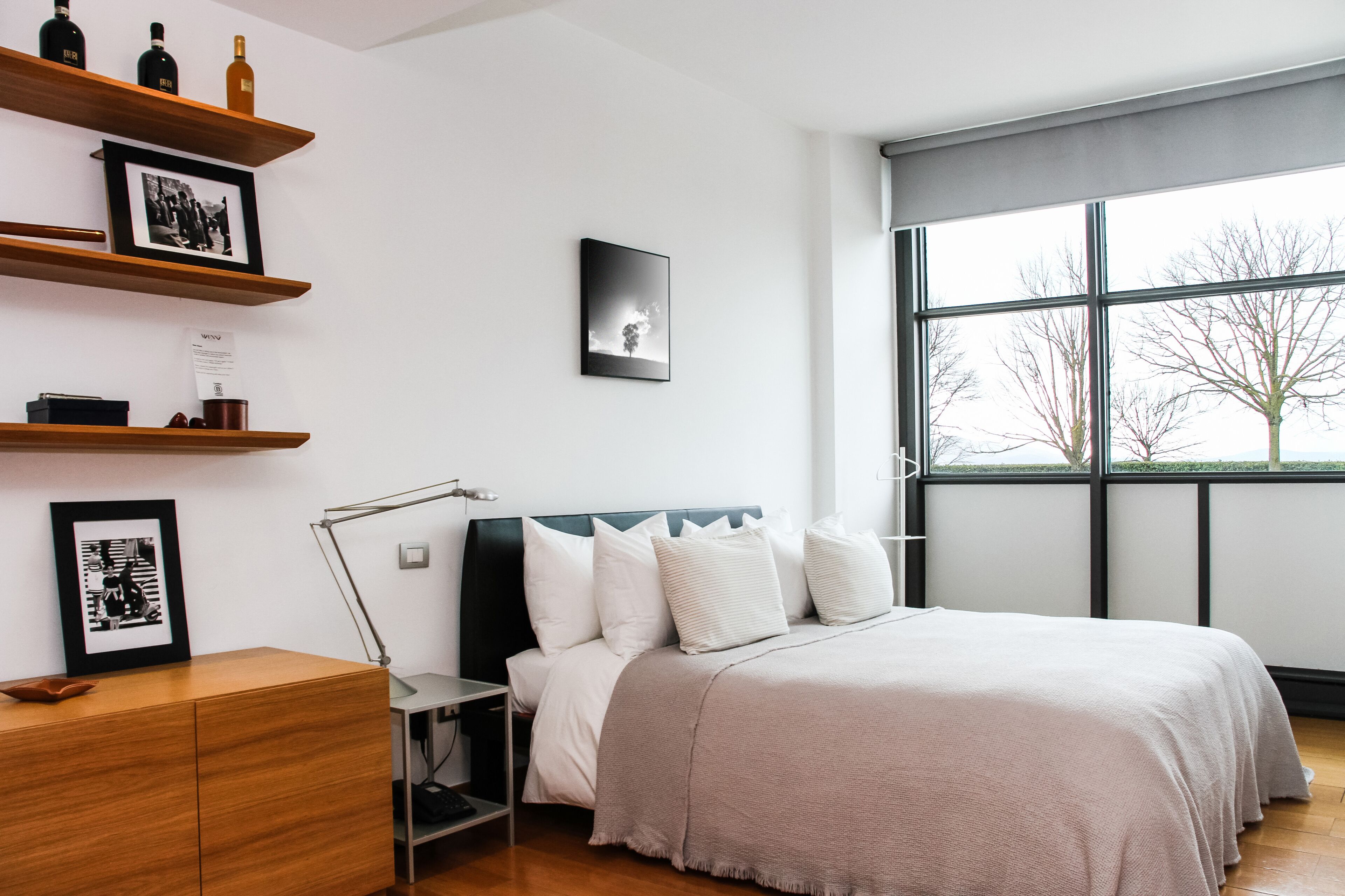 Un dormitorio moderno con un ventanal grande, una cama con cabecera y estantes de madera con fotos enmarcadas y botellas.