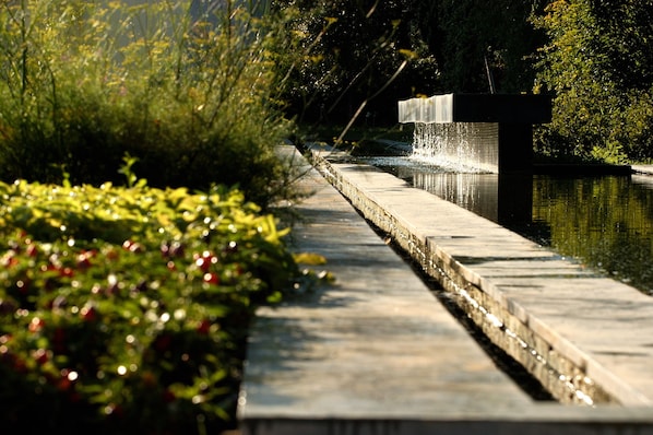 A concrete pathway beside a body of water with a waterfall feature, surrounded by greenery. - Borgo San Gregorio (Sorbo Serpico)