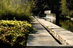 A concrete pathway beside a body of water with a waterfall feature, surrounded by greenery.