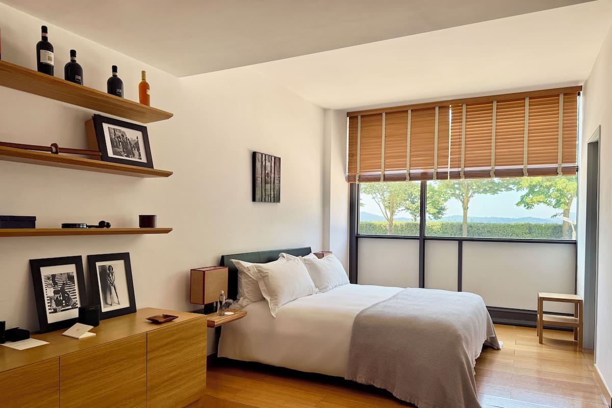 A modern bedroom with a large bed, wooden floor, and a view of trees through a window with blinds.