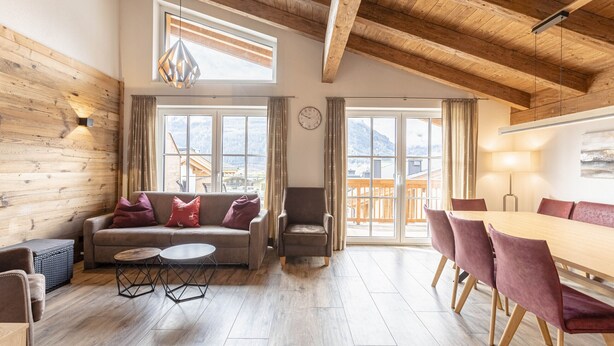 A spacious living area with a wooden ceiling, a sofa with red cushions, a dining table with chairs, and large windows offering a view of mountains. - Superior Chalet # 4C with sauna (Uttendorf)