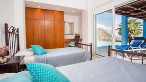 A room with two beds, a wooden wardrobe, a desk, and a view of a pool and mountains. - About Villa Selene (Archangelos)