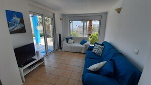 A living room with a TV, a blue sofa, and a white couch. There is a large window with a view of the outdoors. - About Villa Selene (Archangelos)