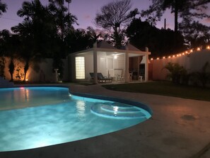 Une piscine à bord incurvé, un patio couvert avec des chaises longues et des guirlandes lumineuses.