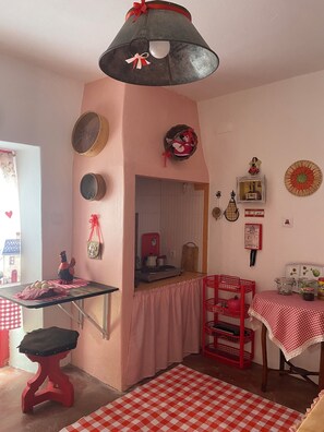 A kitchen with a red and white checkered tablecloth, a red stool, a hanging light fixture, and a wall-mounted shelf with kitchen items. - CASAL DOS CARPINTEIROS 1 - Beach and Countryside (Silveira)