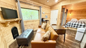 Ein gemütliches Blockhaussinnere mit einer Ledersofa, einem kleinen Tisch, einem an der Wand montierten Fernseher und einem Bett mit einer Karodecke.