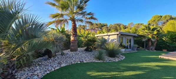 A well-maintained garden with palm trees, a small building, and a clear sky.