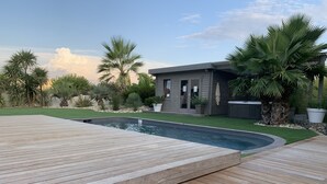 A swimming pool with a wooden deck, surrounded by palm trees and a small building with a patio. - Villa with pool in a haven of peace (Sommières)