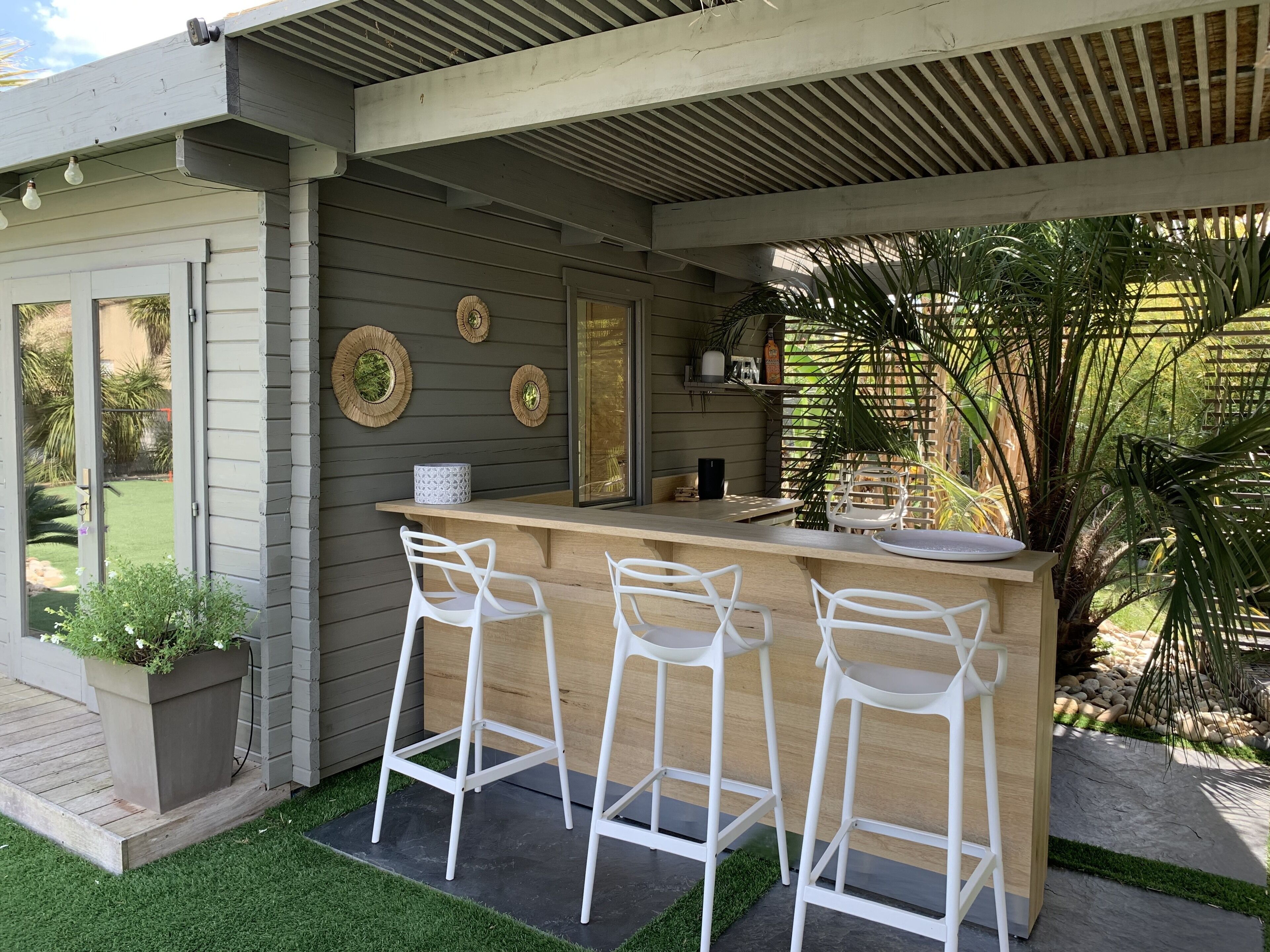 A modern outdoor bar area with a wooden counter, three white bar stools, and decorative wall art.