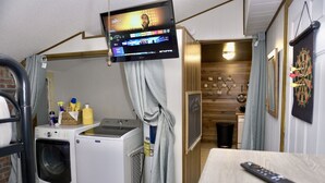A laundry room with a washing machine, dryer, and ironing board. A television is mounted on the wall, and a remote control is on the table. - Cozy Home w/ Doggy Fence (Warsaw)