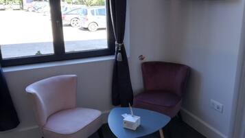 Una habitación con un ventanal grande, una silla rosa, una silla morada y una mesita con un objeto blanco encima.