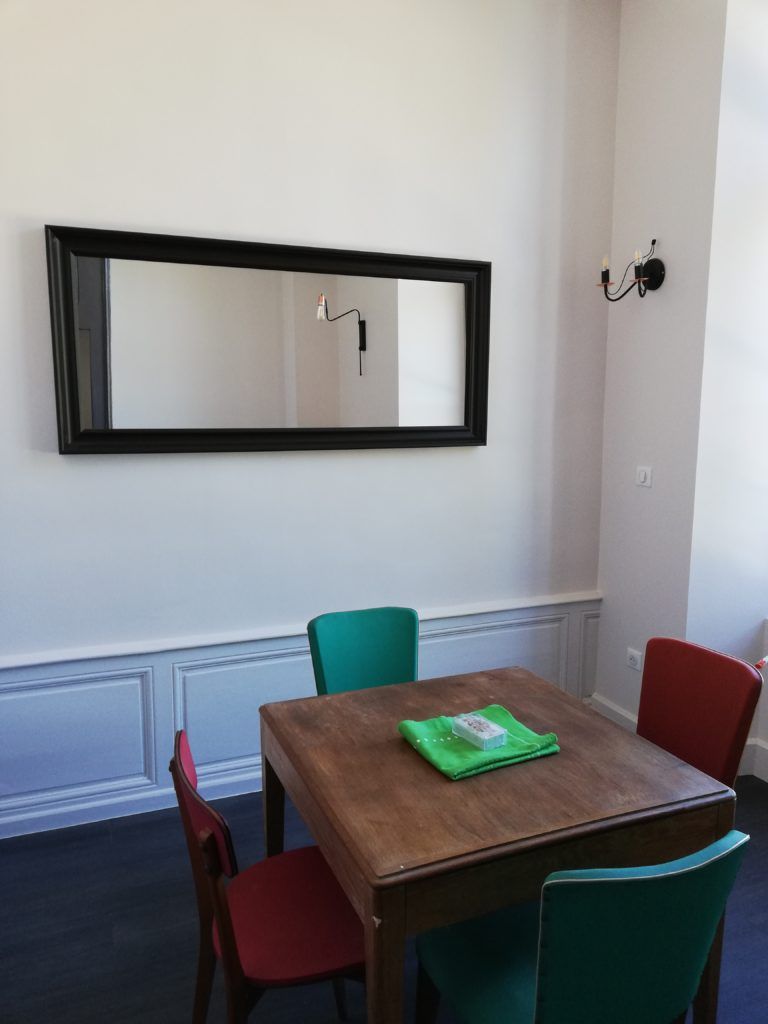 Un coin repas avec une table en bois et quatre chaises, un grand miroir au mur et un luminaire fixé au mur.