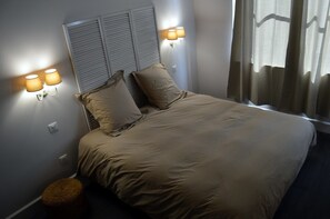 A neatly made bed with a headboard, two wall-mounted lamps, and a small wicker stool. -  Cote Green (Combles-dn-Barrois)