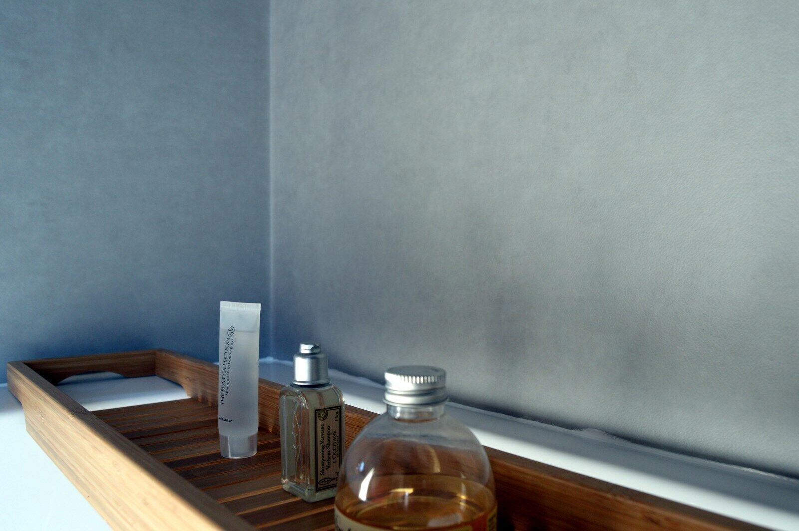 Ein Holztablett mit einer Shampooflasche, einer Lotionflasche und einer Ölflasche auf einer weißen Oberfläche vor einer blauen Wand.