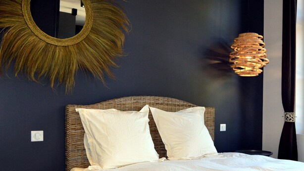 Une chambre à coucher avec une tête de lit en osier, du linge de lit blanc, un miroir rond et un luminaire suspendu original.