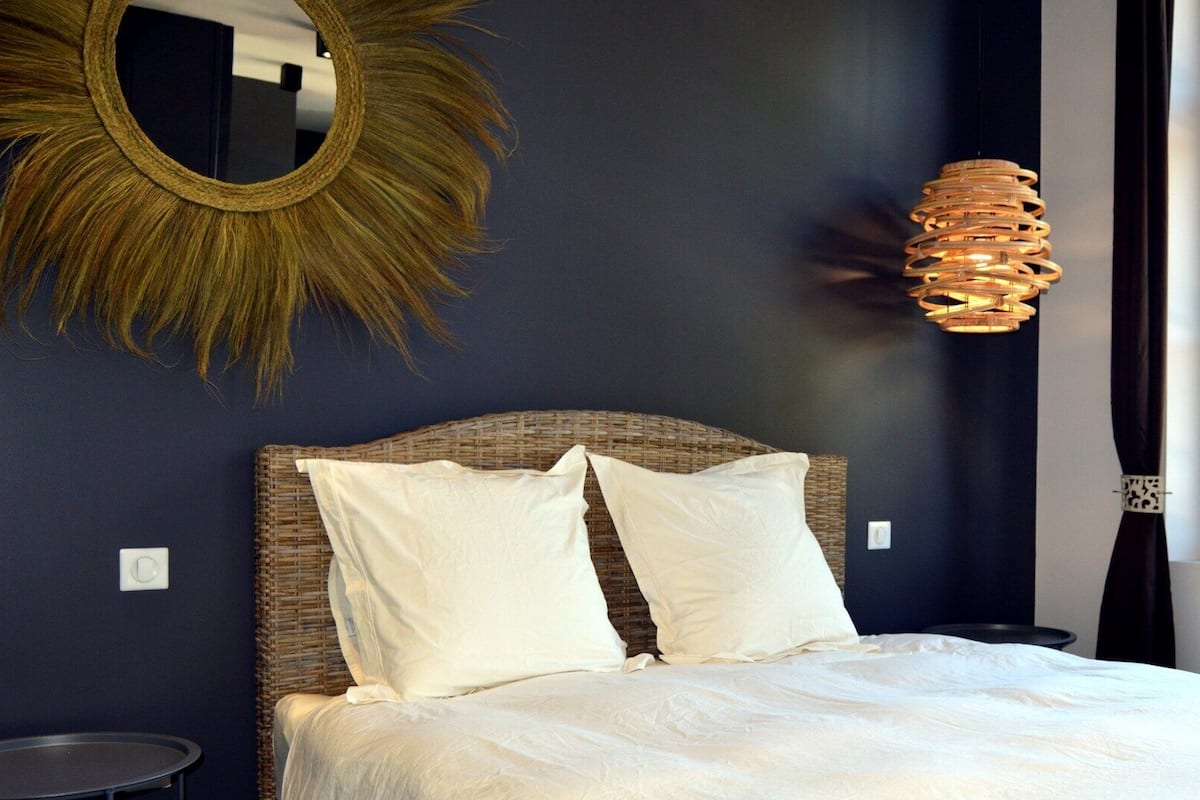 Un dormitorio con cabecera de mimbre, ropa de cama blanca, un espejo redondo y una lámpara colgante original.