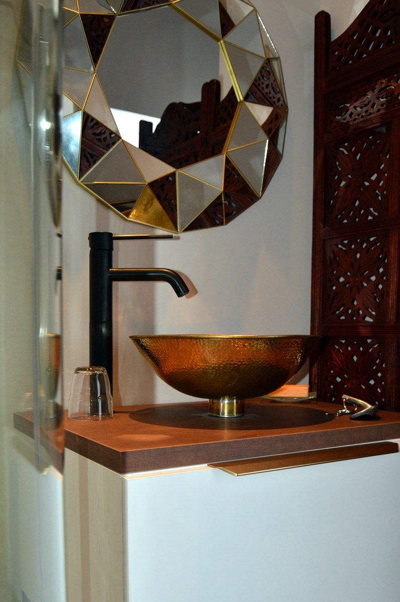 Une salle de bain avec un miroir aux formes géométriques, un lavabo en cuivre et un robinet noir.
