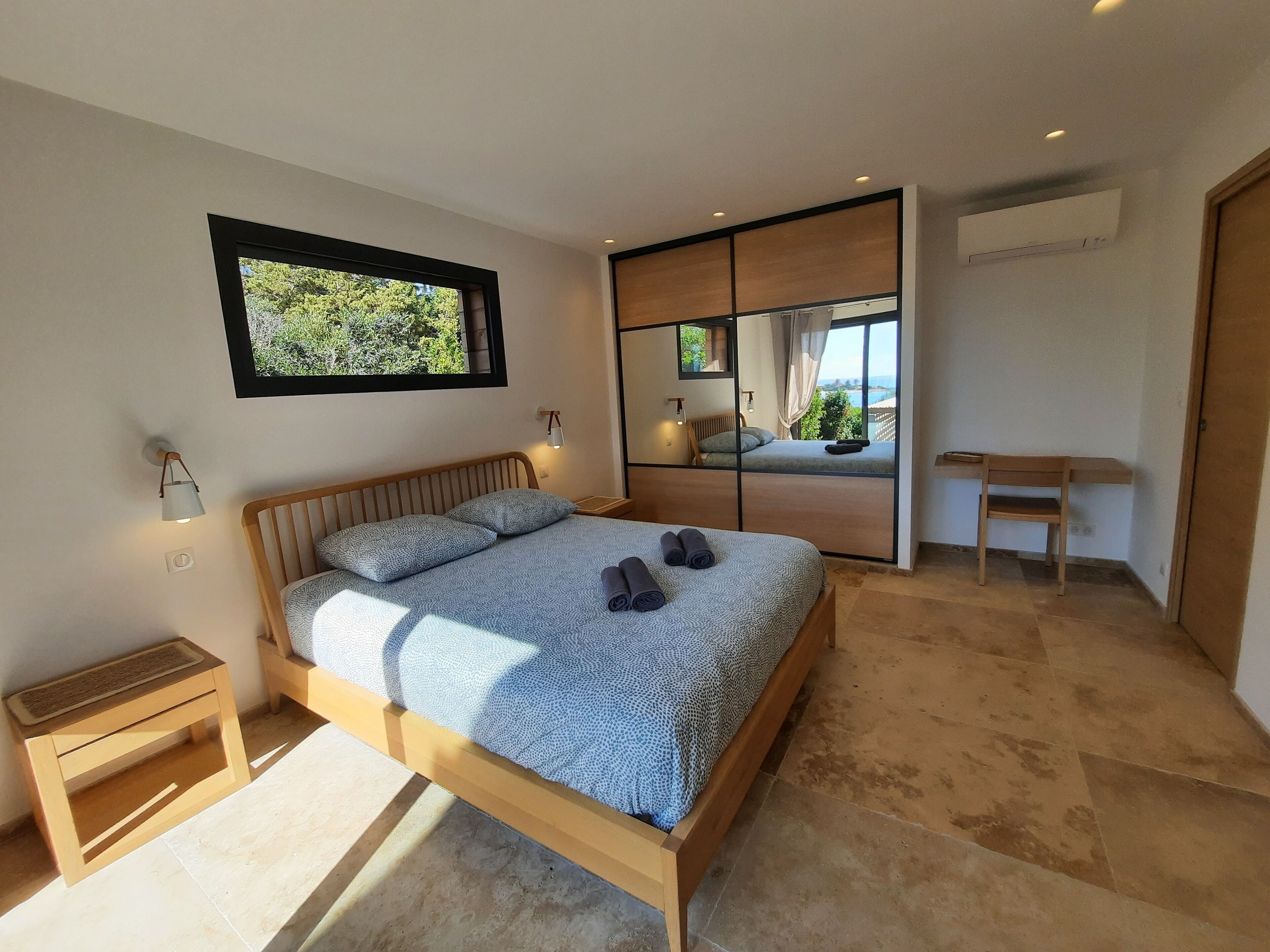 A modern bedroom with a large bed, a bedside table, a small desk, and a view through a sliding door.