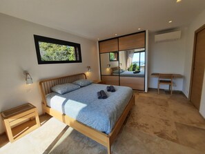 A modern bedroom with a large bed, a bedside table, a small desk, and a view through a sliding door. - Beachfront villa. (Sartène)