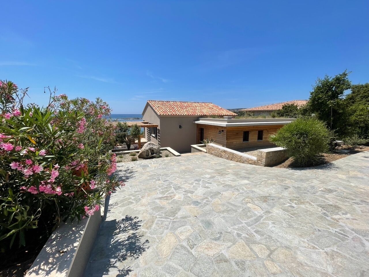 A modern house with a stone patio, a small garden with pink flowers, and a view of the sea.
