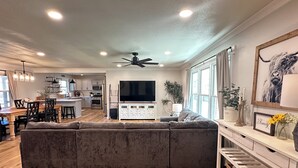 Una sala moderna con un sofá seccional gris grande, un comedor con mesa y sillas de madera, una cocina con armarios blancos y un televisor grande, y un ventilador de techo.