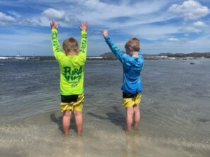 Two children standing in shallow water with their arms raised, wearing long-sleeve shirts and shorts. - Private 2BR Villa, Resort Pool, Walkable to Beach (Tamarindo)