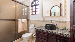 A bathroom with a glass shower enclosure, a toilet, a vanity with a stone-patterned countertop, and a mirror with decorative lights.