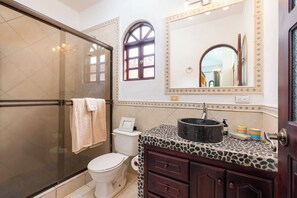 Een badkamer met een glazen douchecabine, toilet, wastafel met een aanrechtblad met steenmotief en een spiegel met decoratieve verlichting.