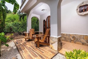A covered outdoor seating area with wooden chairs and a table, surrounded by greenery and a stone wall. - Private 2BR Villa, Resort Pool, Walkable to Beach (Tamarindo)