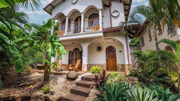 A two-story building with a balcony, surrounded by tropical vegetation.