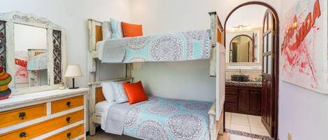 Een stapelbed met beddengoed met een patroon, een dressoir met lades, een spiegel en een zichtbare badkamer door een boogvormige doorgang.