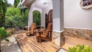 A covered outdoor seating area with wooden chairs and a table, surrounded by greenery and a stone wall. - Private 2BR Villa, Resort Pool, Walkable to Beach (Tamarindo)