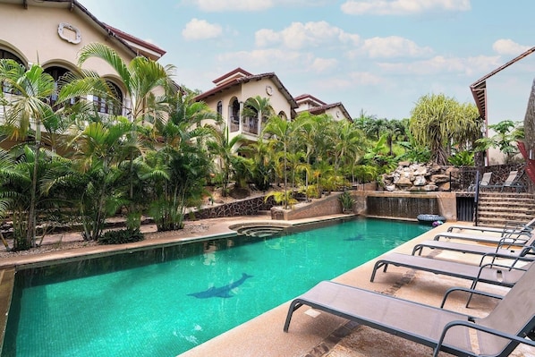 A swimming pool with lounge chairs, surrounded by tropical plants and a building with arched windows.