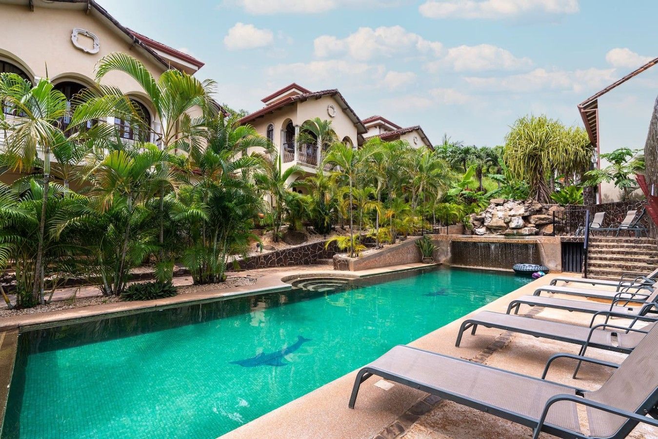 Piscina com espreguiçadeiras, rodeada por plantas tropicais e um edifício com janelas em arco.
