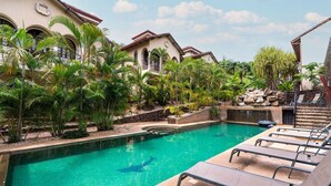 A swimming pool with lounge chairs, surrounded by tropical plants and a building with arched windows.
