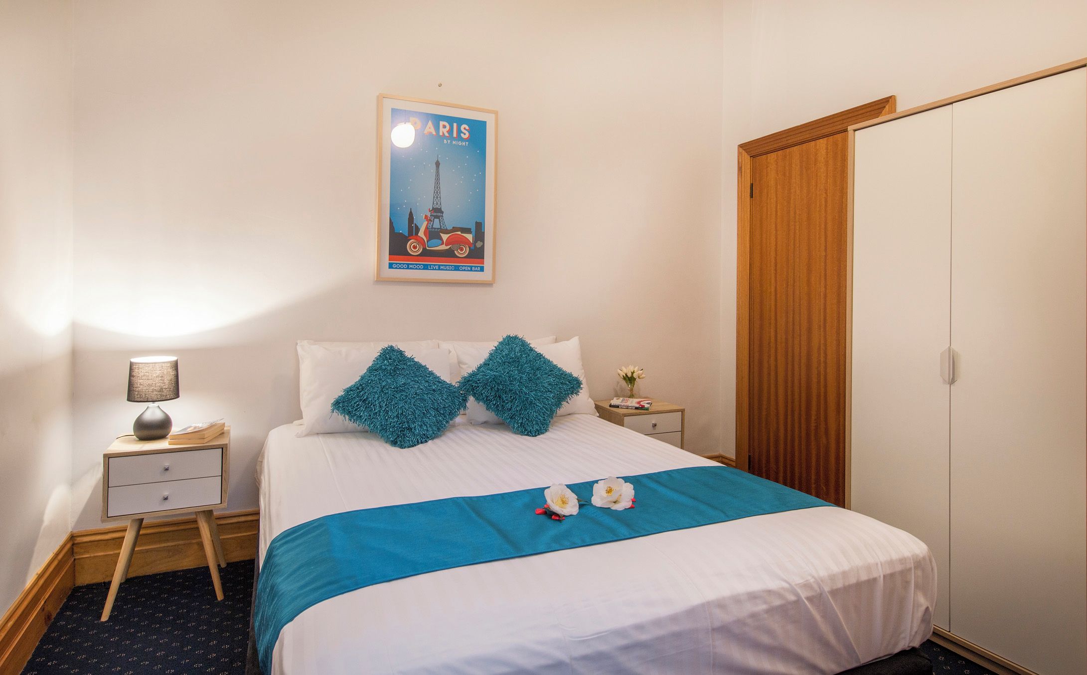 A neatly arranged bedroom with a bed, a nightstand, a lamp, a framed poster, and a wardrobe.