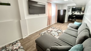 Una sala de estar moderna con un sofá gris, un televisor de pantalla plana montado en la pared, una zona de cocina con un refrigerador y una mesa de comedor con sillas.