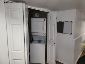 A compact laundry room with a washer and dryer, a small shelf with a whiteboard, and a door leading to another room.