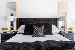 A bedroom with a tufted headboard, two bedside tables with lamps, a large mirror, and a black fur throw.