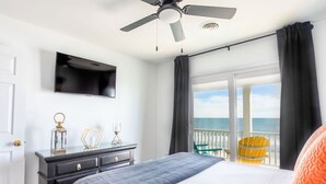 Et hotelværelse med seng, ventilator, fjernsyn ophængt på væggen, kommode og balkon med havudsigt.