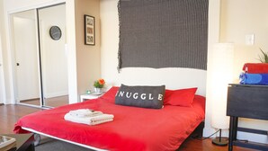 En snyggt bäddad säng med ett rött överkast och en kudde med ordet ”NUGGLE” på. Bredvid sängen står ett litet bord med en lampa och en växt.