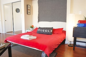 En pænt redt seng med et rødt sengetæppe og en pude med ordet "NUGGLE" på. Der er et lille bord med en lampe og en plante ved siden af sengen.