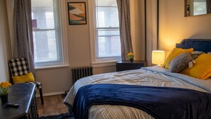 Un dormitorio con cama, mesita de noche, silla y ventanas con cortinas.