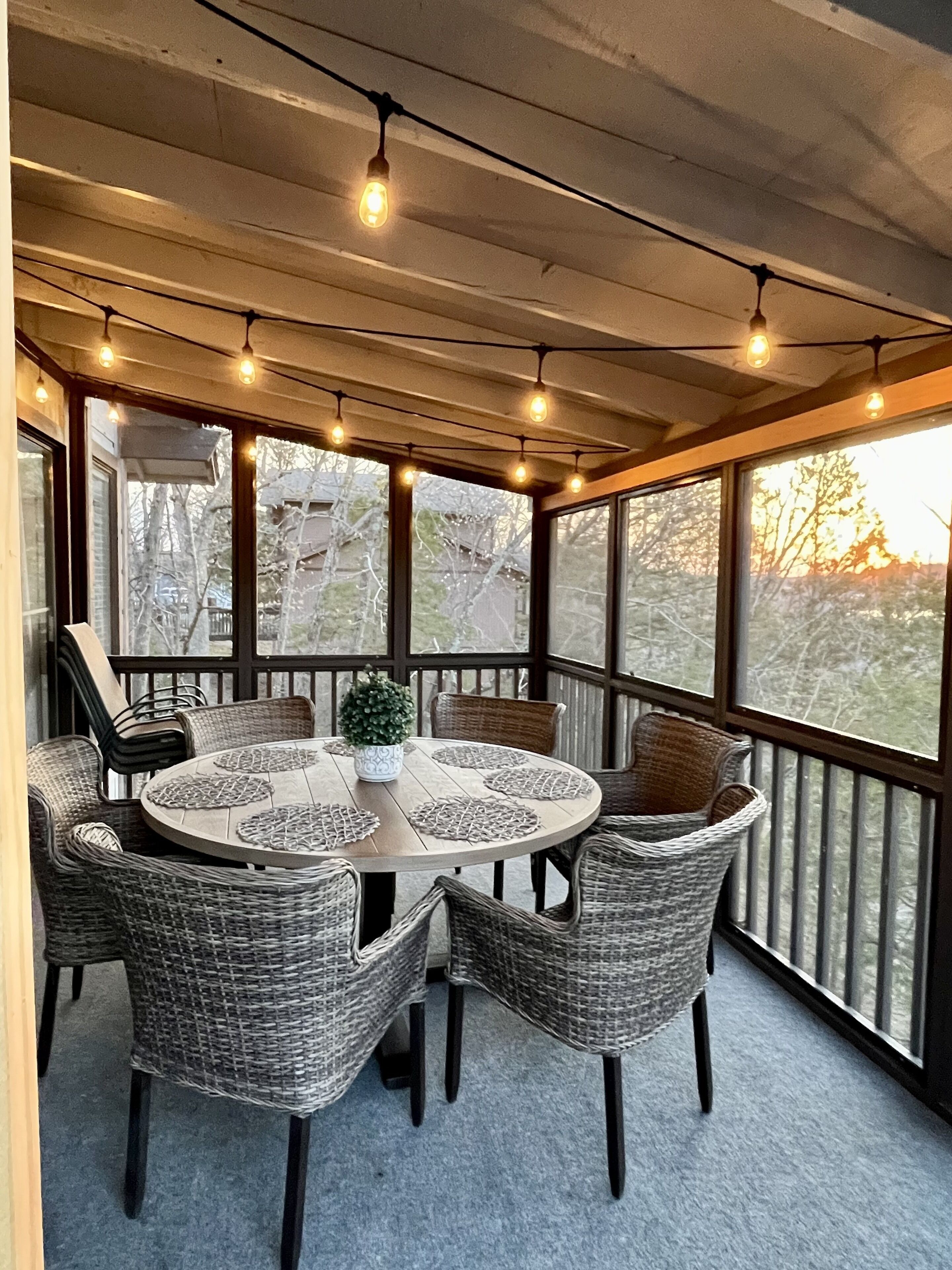 Un área de comedor al aire libre techada con sillas de mimbre y una mesa redonda, luces colgantes y ventanas amplias que ofrecen vista a árboles y al atardecer.
