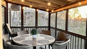 Uma área de refeições ao ar livre coberta, com cadeiras de vime e uma mesa redonda, luzes de festa penduradas e grandes janelas com vista para árvores e um pôr do sol.