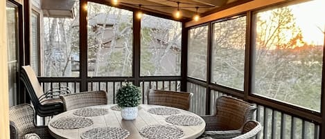 Un área de comedor al aire libre techada con sillas de mimbre y una mesa redonda, luces colgantes y ventanas amplias que ofrecen vista a árboles y al atardecer.