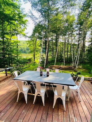 An outdoor deck with a rectangular table and white chairs, surrounded by a forested area. - Lakefront Serenity in the Catskills near All Star Village (Franklin)