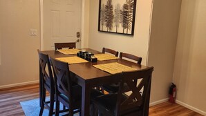 Un espace repas comprenant une table en bois, des chaises, un tapis à motifs, un luminaire suspendu au plafond et une plante fixée au mur.